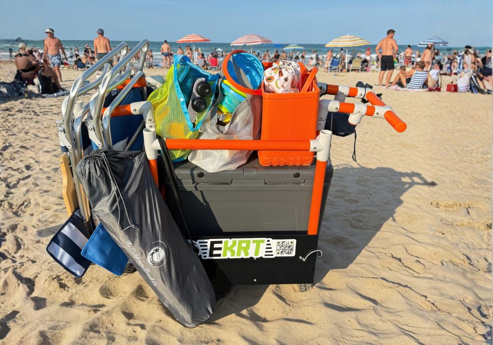 Orange e-KRT foldable electric beach cart with balloon sand tires, loaded with a cooler, chairs, and beach bag on a white background