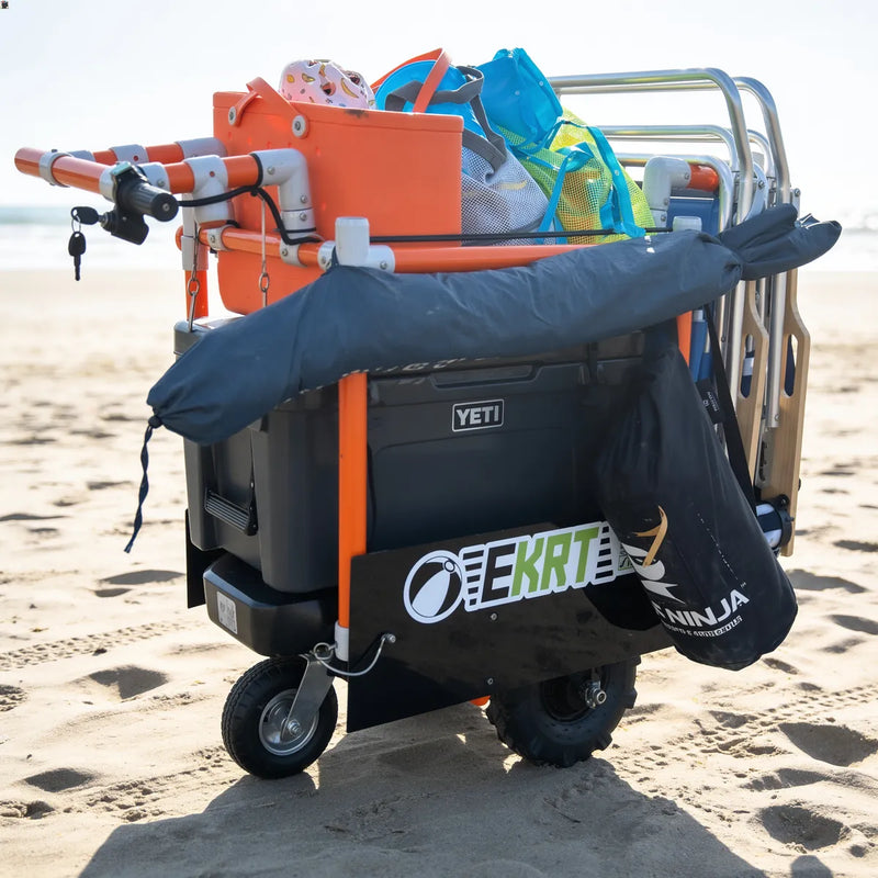 An e-KRT electric beach cart loaded with gear, including an orange storage box, a blue bag, and a large black YETI cooler, is parked on sandy beach sand.
