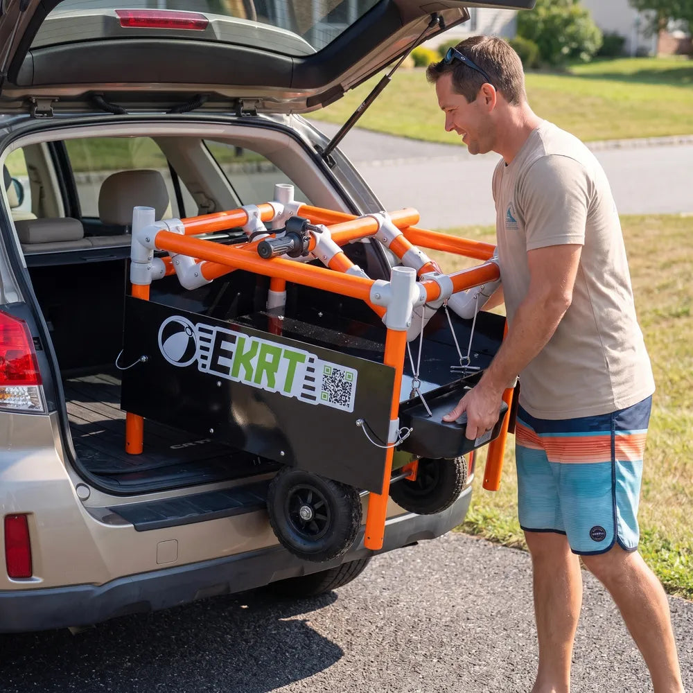 A man is loading a portable black and orange e-KRT electric beach cart into the trunk of an SUV outdoors on a paved driveway.