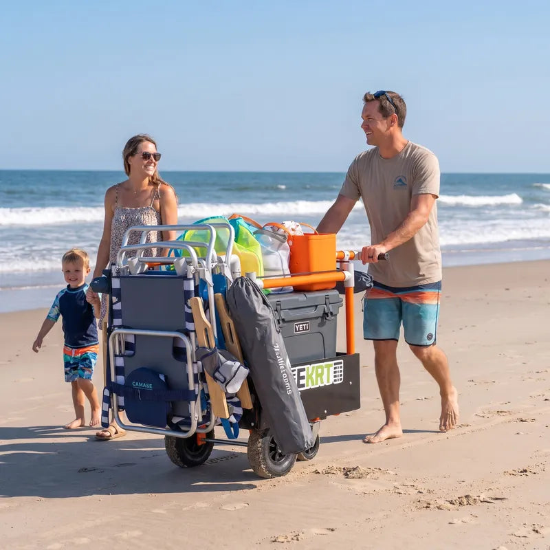 A family of three pushes an e-KRT electric beach cart loaded with folding chairs, coolers, and beach gear on a sandy beach.
