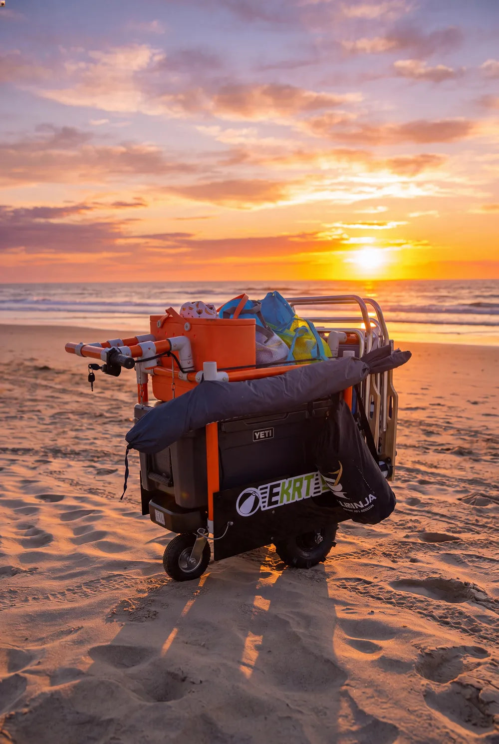 A black and orange beach e-KRTloaded with gear, parked on a sandy beach at sunset with the ocean and sky in the background.