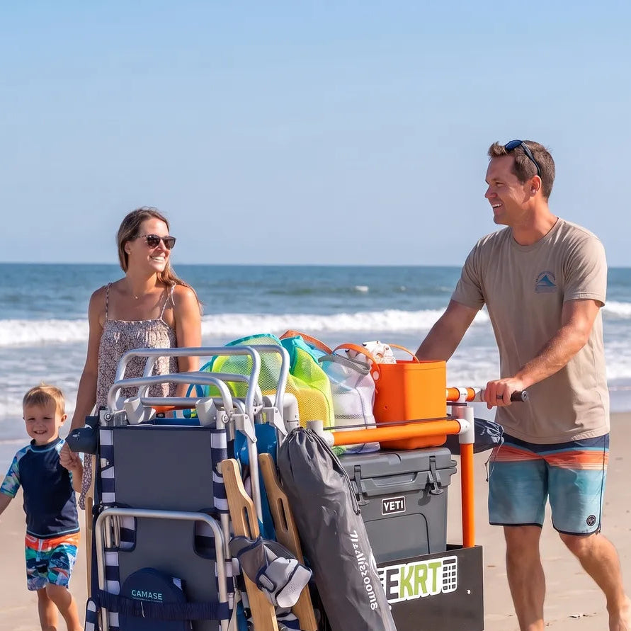 man pushing an orange e-KRT foldable electric beach cart loaded with chairs and a cooler across soft sand toward the ocean